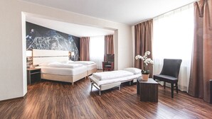 Hotellihuoneessa on suuri sänky, pienempi sohva, työpöytä ja tuoli. Seinällä on taulu ja ikkunassa verhot.