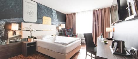 A hotel room with a bed, bedside table, lamp, TV, desk, chair, and a large framed picture on the wall.