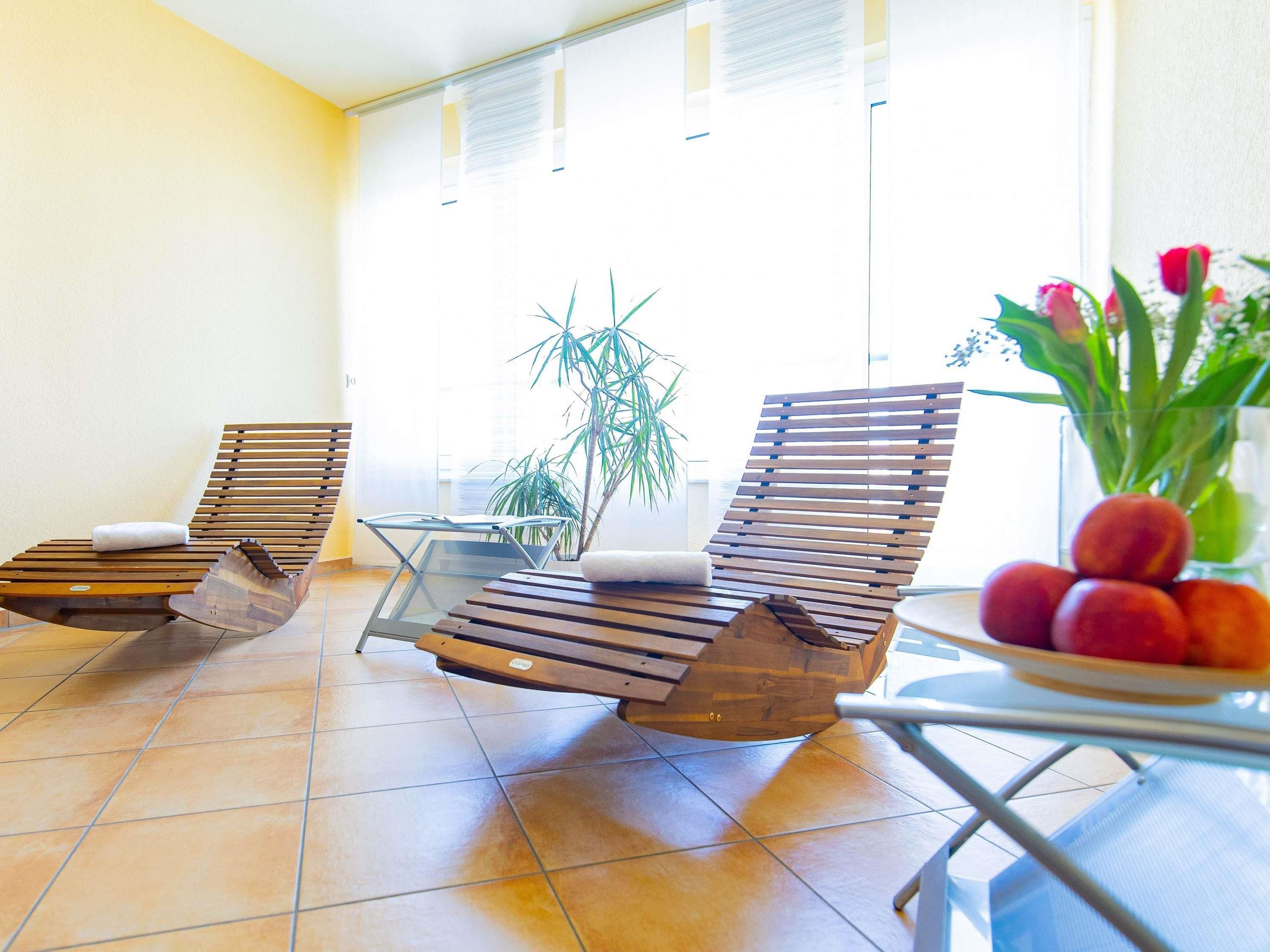 Una habitación iluminada por el sol con dos sillones de madera, una mesa de vidrio, una planta en maceta y un florero con flores.