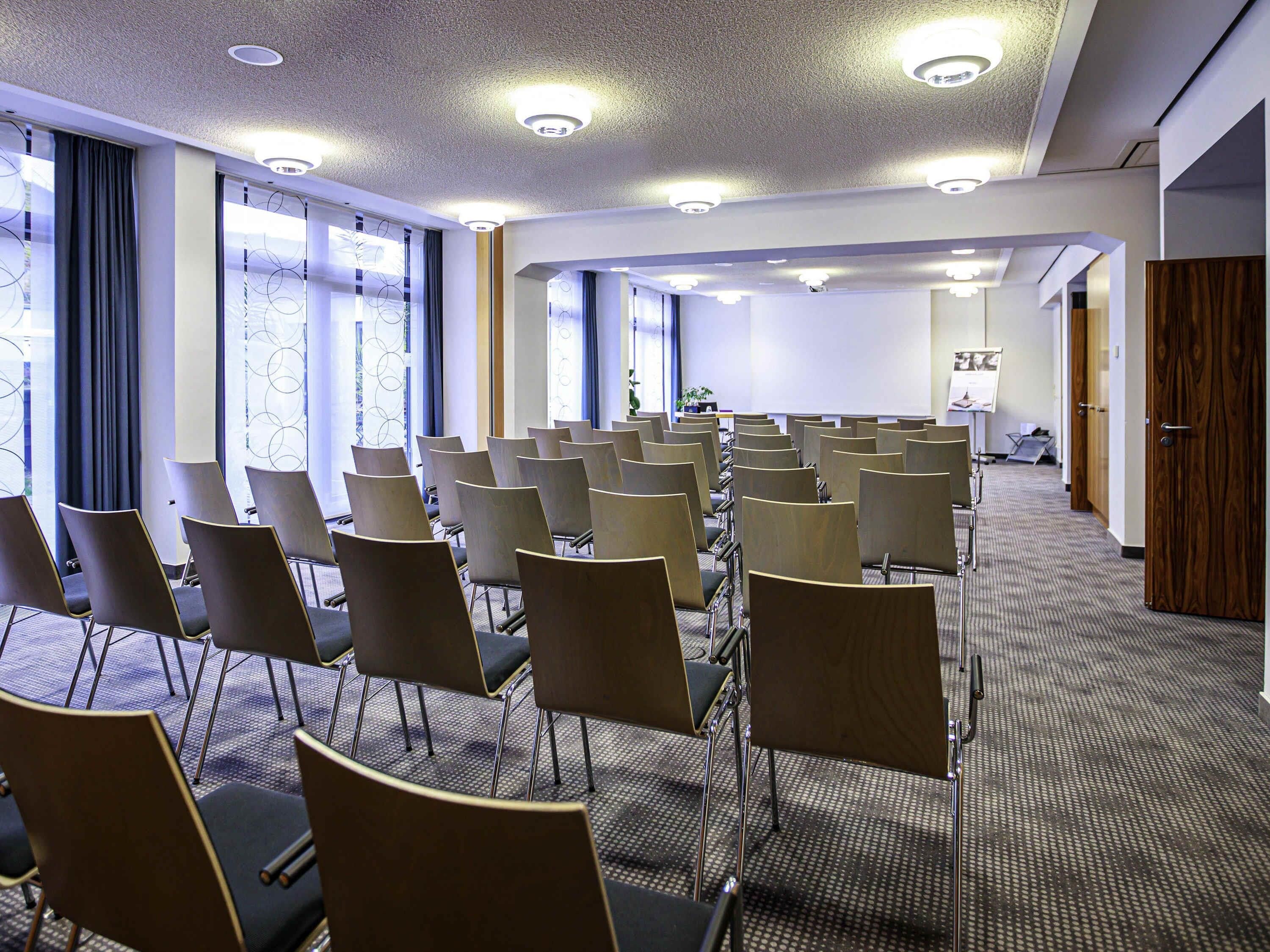 Un salón de conferencias con filas de sillas, una pantalla de proyector y una zona para presentaciones.