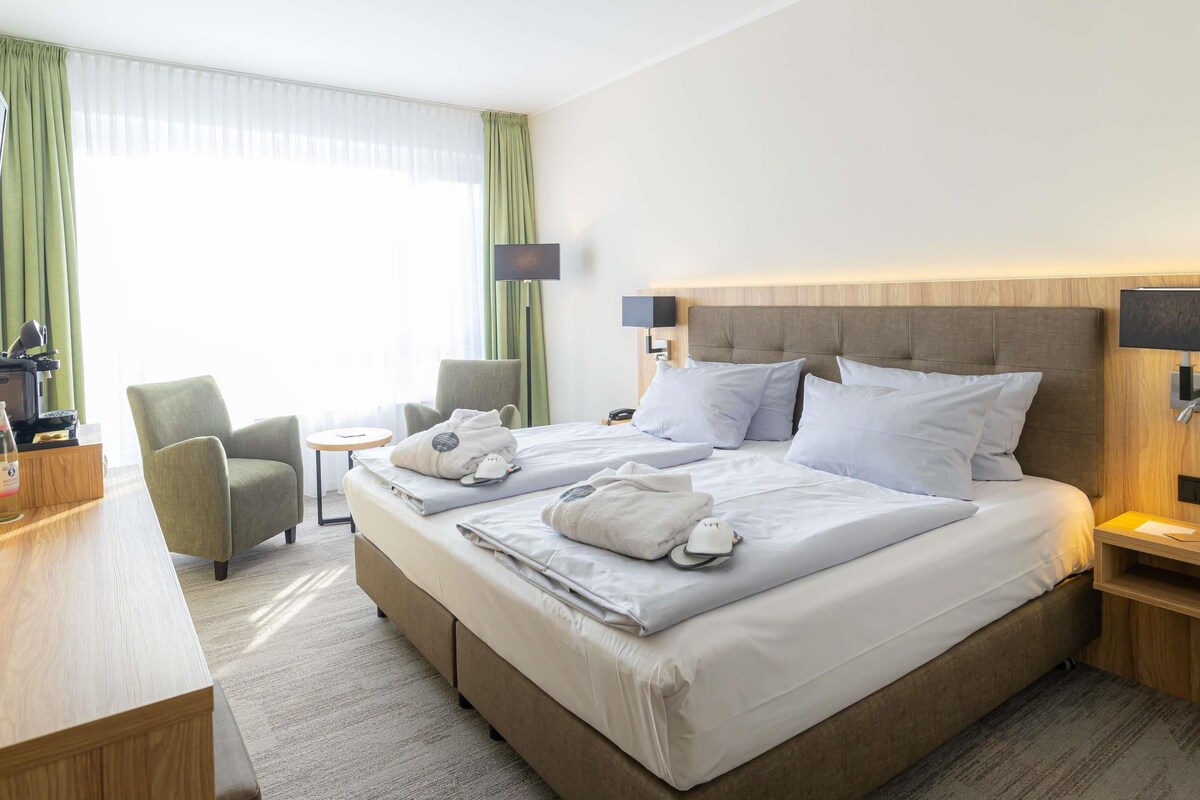 A hotel room with a large bed, a desk, a chair, a small table, and a lamp.