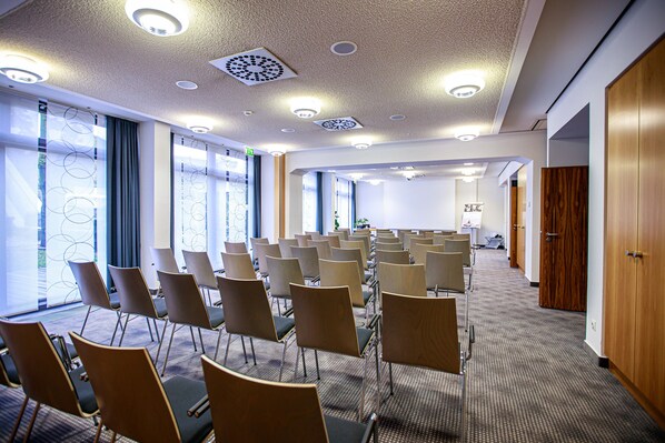 Une salle de conférence avec des rangées de chaises, de grandes fenêtres et un sol recouvert de moquette.