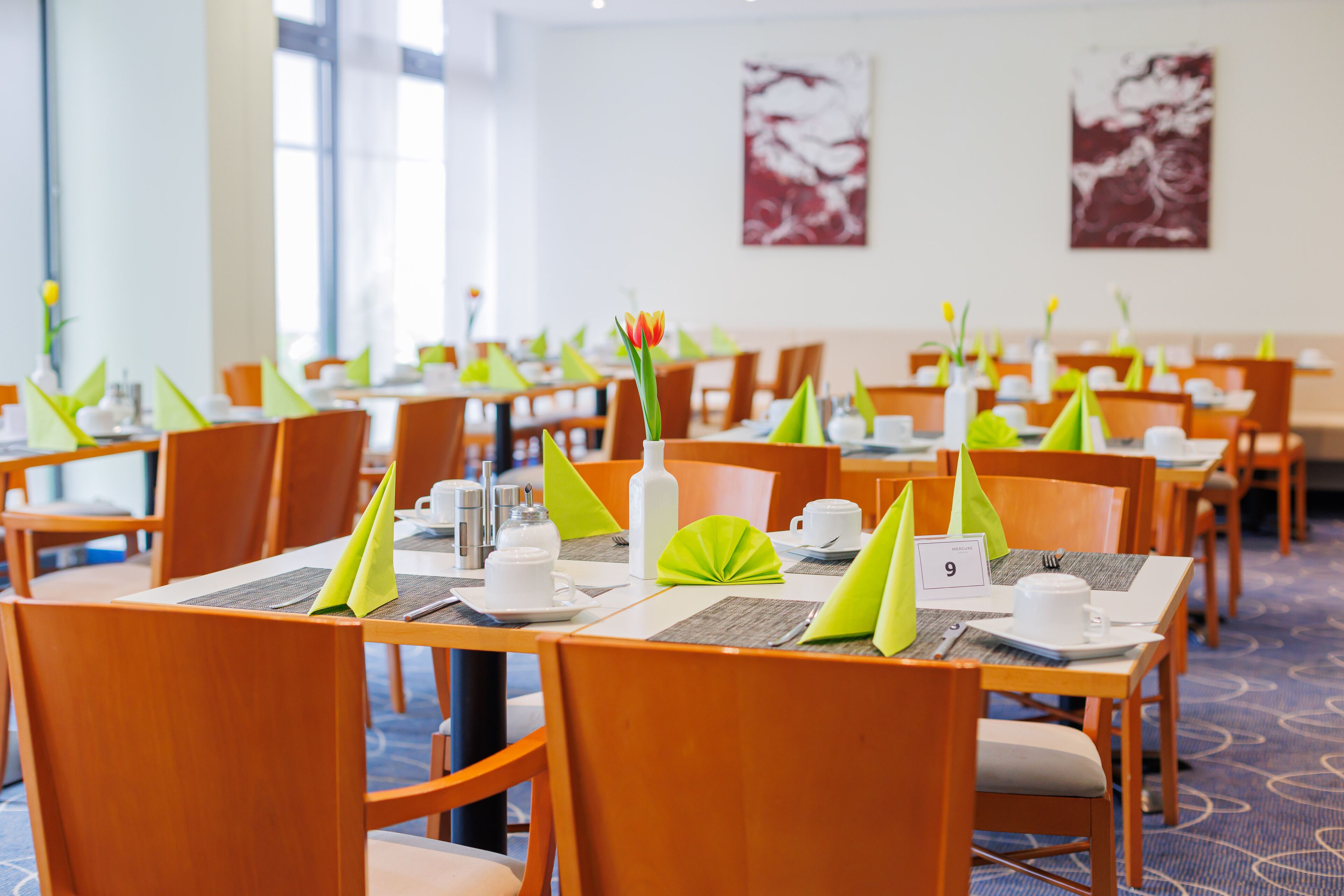 Un comedor de hotel con mesas dispuestas, servilletas verdes, vajilla blanca y posavasos numerados.