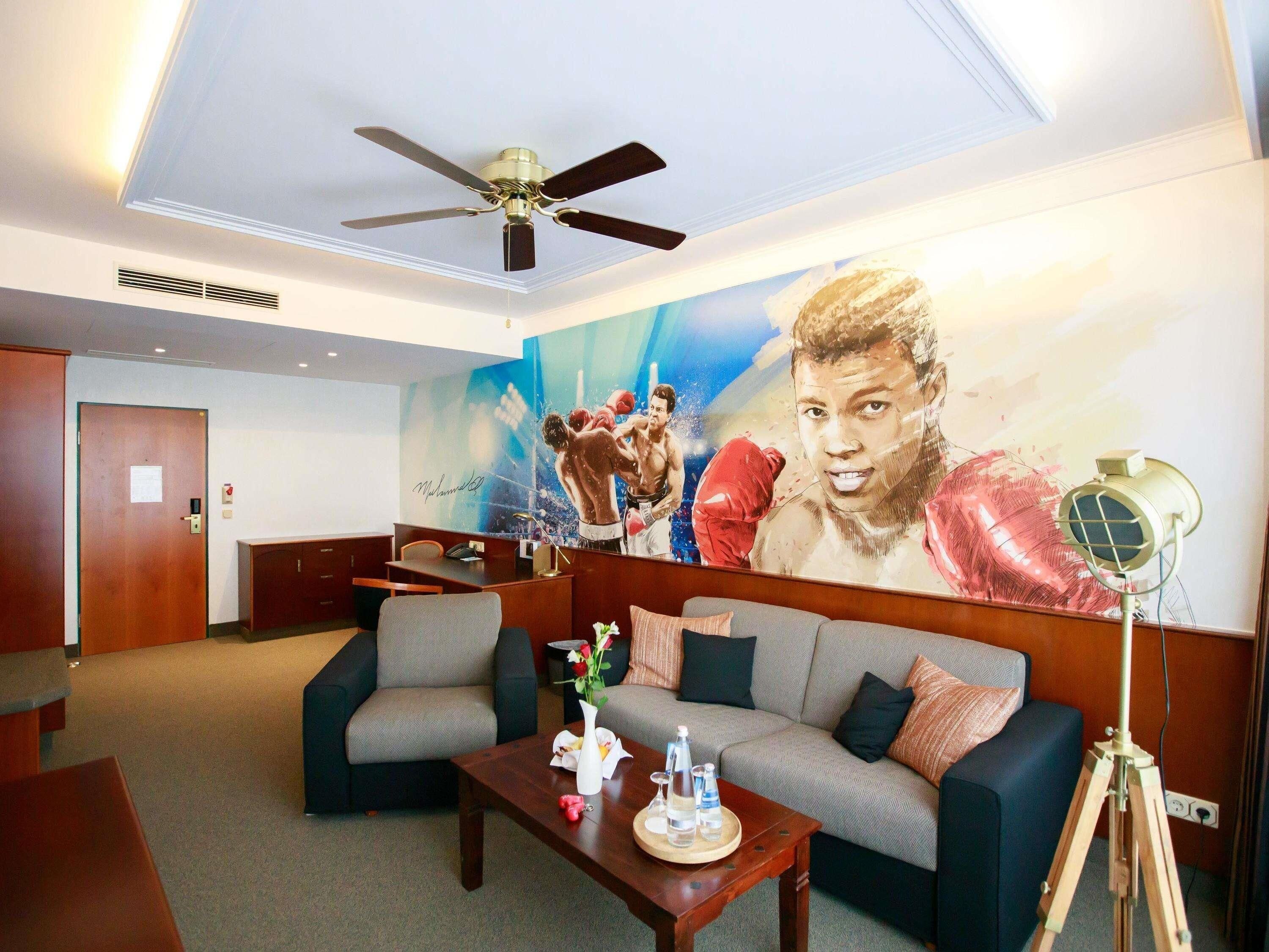 Una hall d'albergo con una ventola a soffitto, un grande murale, un divano, un tavolino da caffè e una lampada con treppiede.