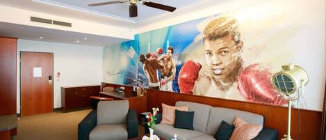 Una hall d'albergo con una ventola a soffitto, un grande murale, un divano, un tavolino da caffè e una lampada con treppiede.