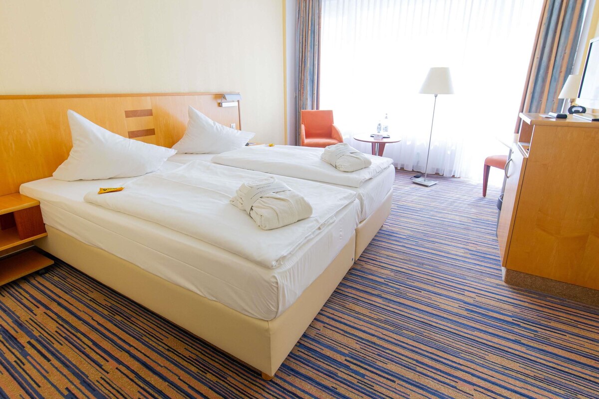 A hotel room with two beds, a desk, a chair, and a lamp.