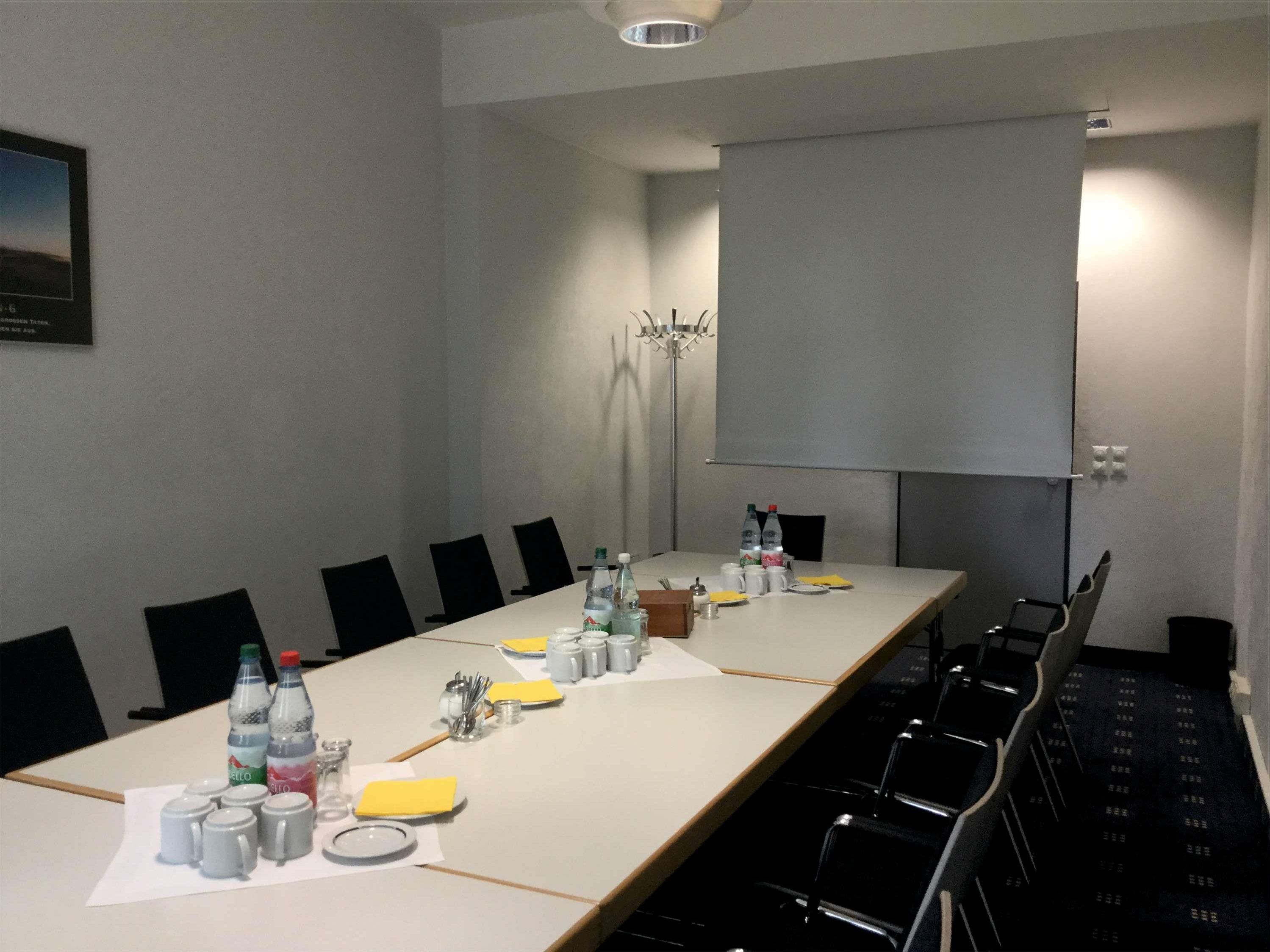 Una sala de conferencias con una mesa larga, sillas, botellas de agua, tazas y una pantalla de proyector.