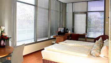A hotel room with a bed, a desk, a chair, a television, and large windows with blinds.
