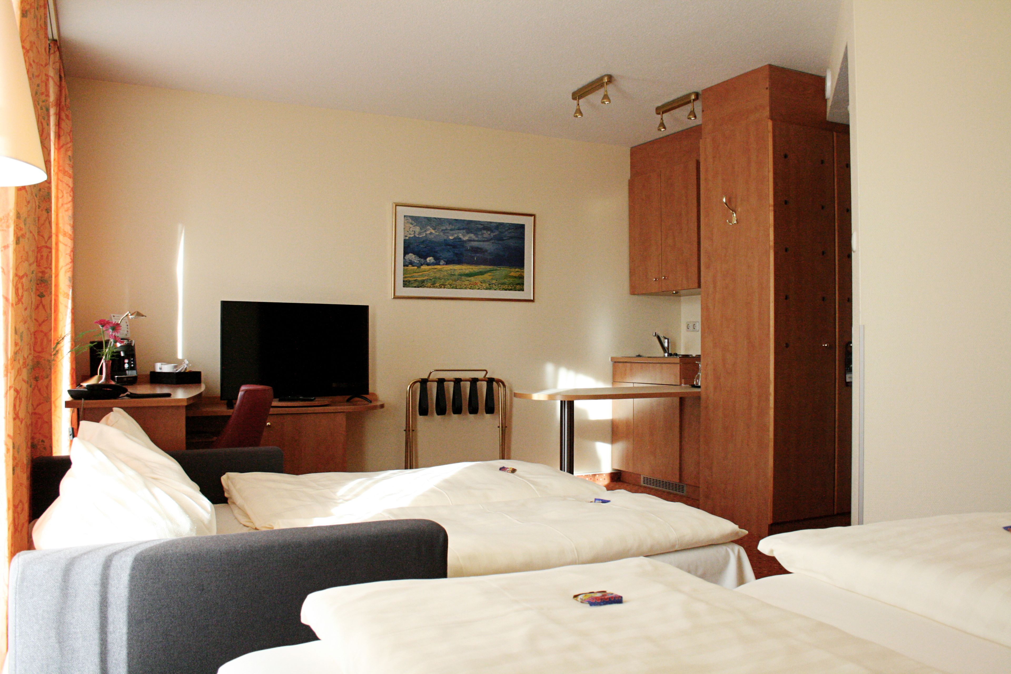 Habitación de hotel con dos camas, un escritorio con televisor, una silla, armario y un cuadro en la pared.