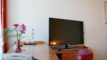 Habitación de hotel con un escritorio de madera, un televisor de pantalla plana, un teléfono, un florero con flores y una botella de agua.