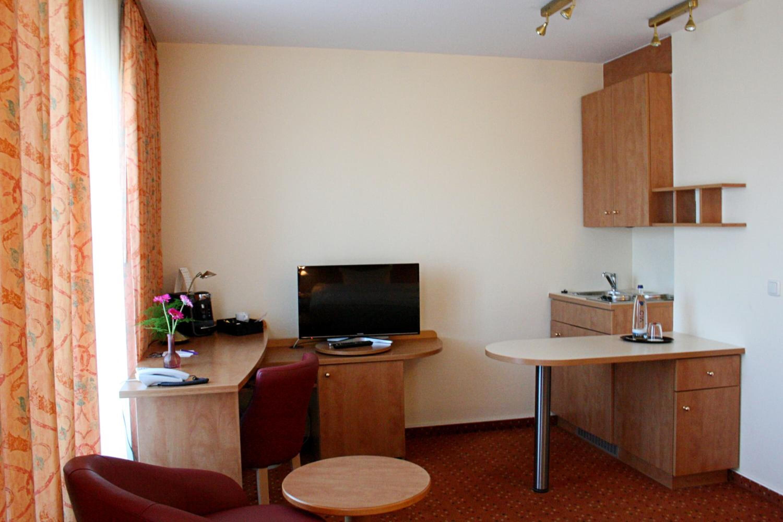Ein kleines Hotelzimmer mit Schreibtisch, Fernseher, Stuhl, rundem Tisch und Küchenzeile mit Spüle und Schrank.