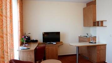 Ein kleines Hotelzimmer mit Schreibtisch, Fernseher, Stuhl, rundem Tisch und Küchenzeile mit Spüle und Schrank.