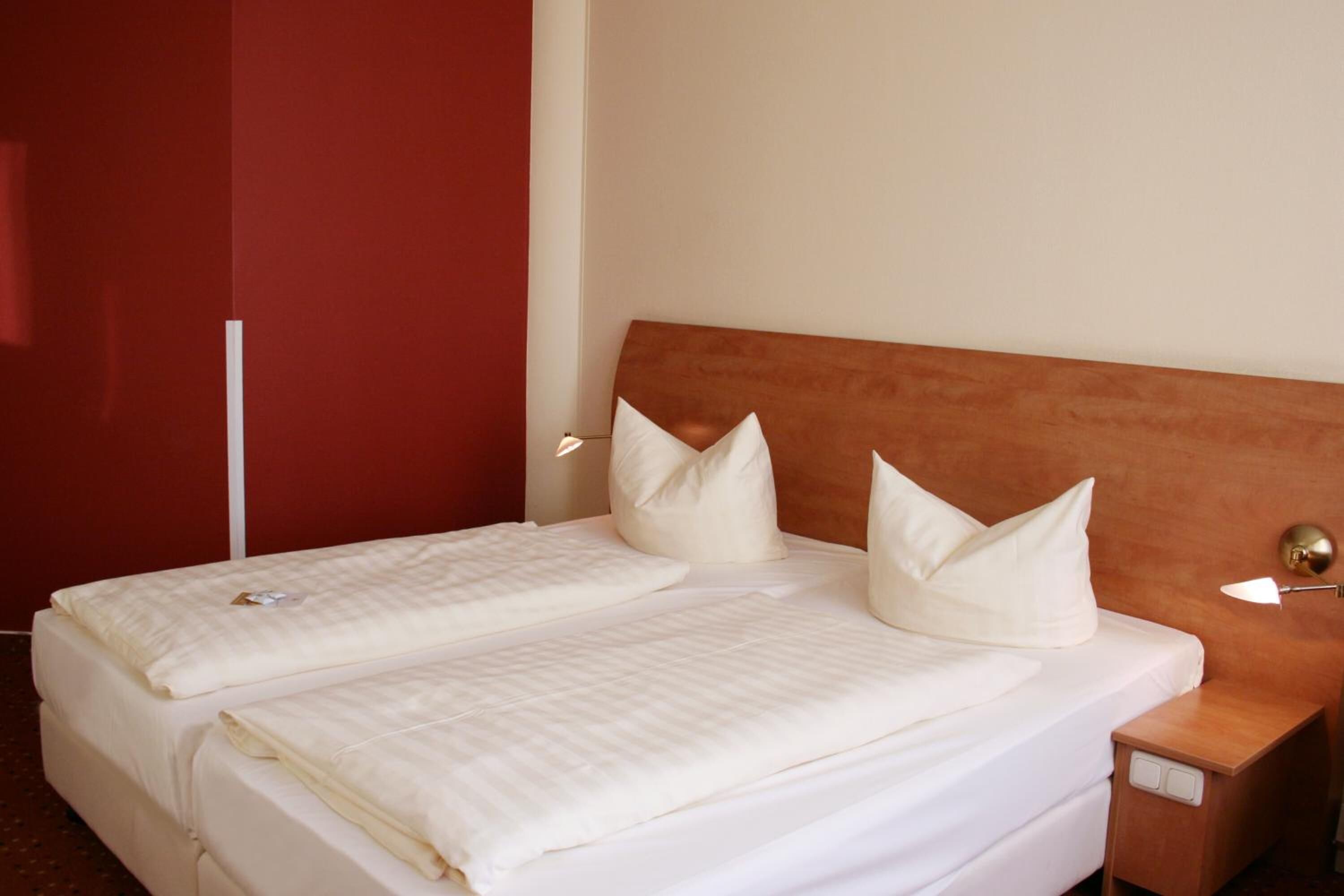 A double bed with white bedding and pillows, a wooden headboard, a bedside table with a lamp, and a red accent wall.