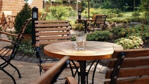 Un patio exterior con mesas y sillas de madera, rodeado de follaje y un edificio de ladrillo.