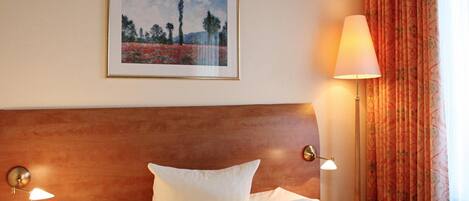 A hotel room with a wooden headboard, a framed landscape painting, a white bed with a pillow, a bedside lamp, and a window with patterned curtains.