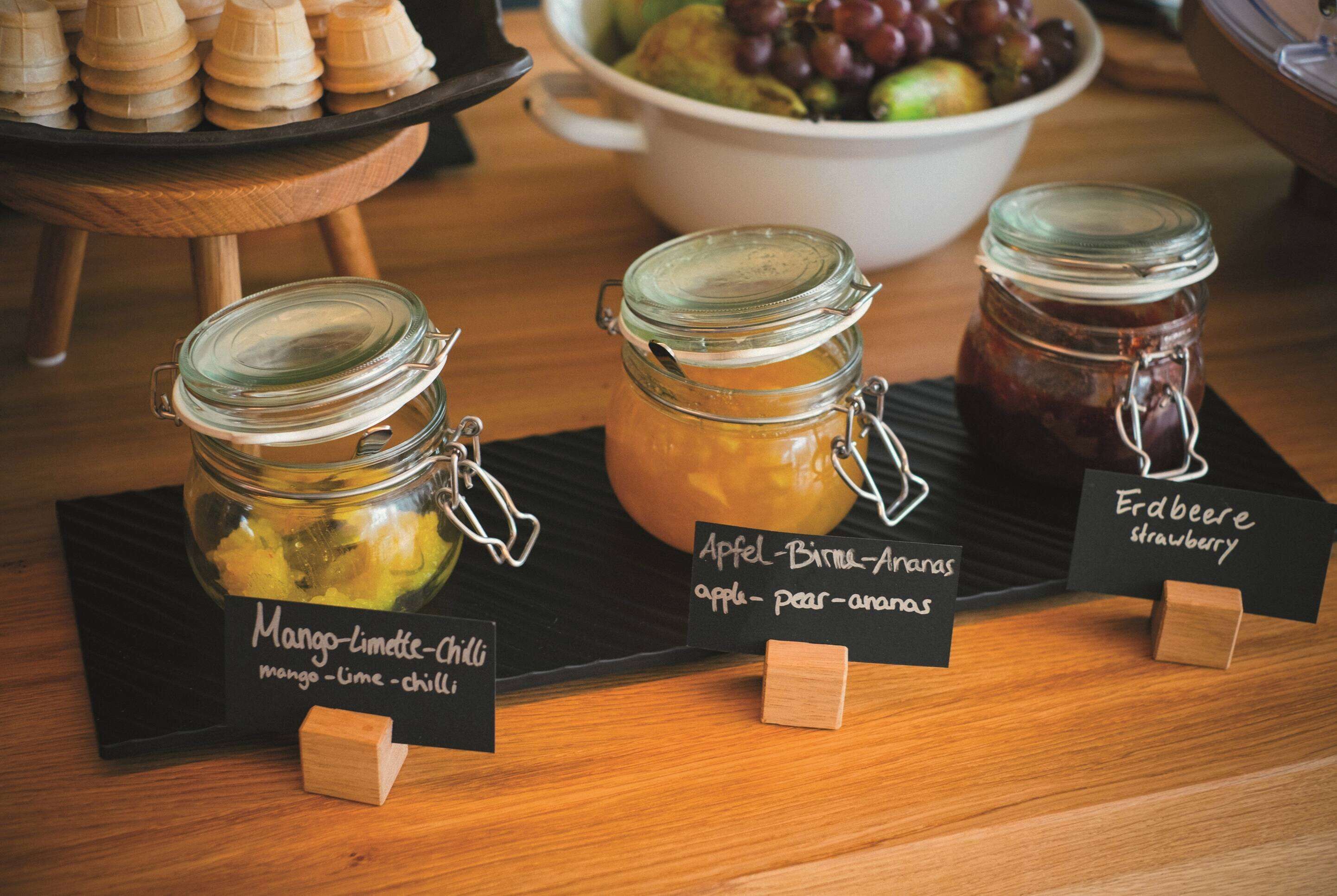 Drei Gläser Konfitüre, beschriftet mit ihren Inhalten: Mango-Limette-Chili, Apfel-Birne-Aronia und Erdbeer-Himbeere.