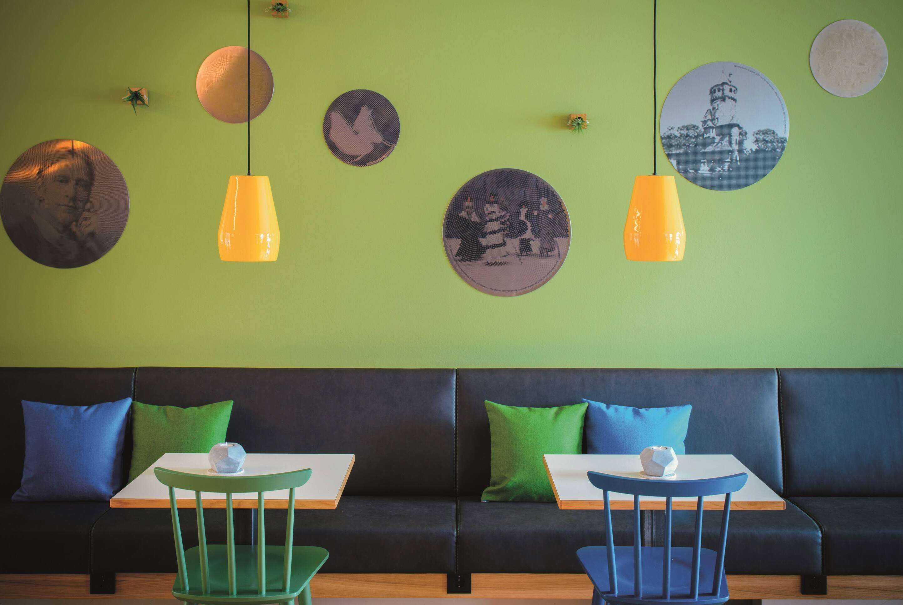 A modern interior with a green wall, two black sofas, and two white tables with blue chairs.