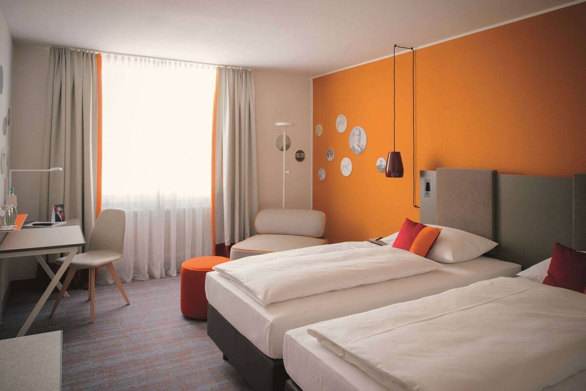 A hotel room with two beds, a desk, a chair, a lamp, and a clock on the wall.