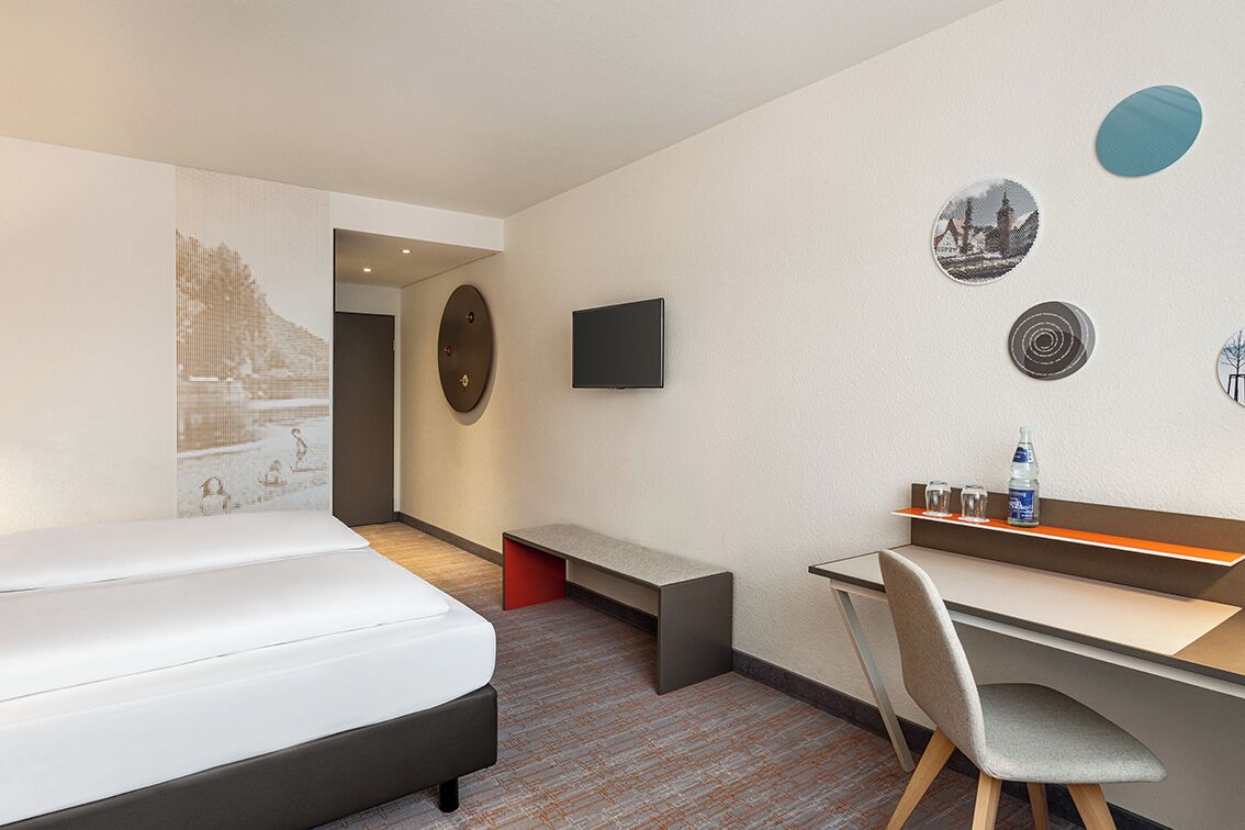 Ett hotellrum med en säng, ett skrivbord och en tv. Det finns dekorationer på väggarna och en bänk.