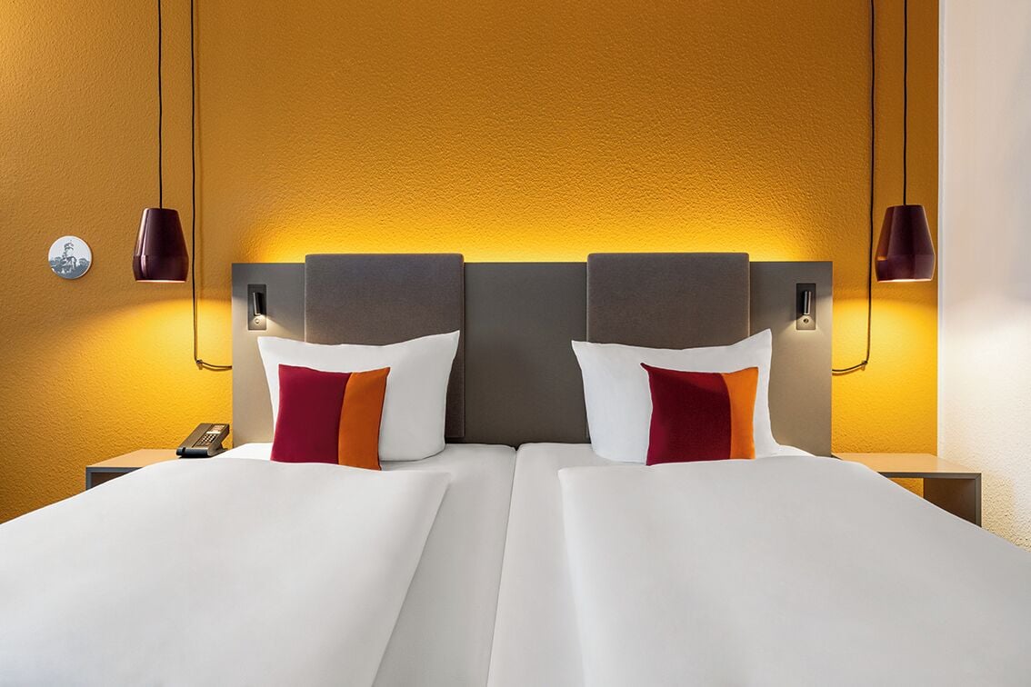 Un lit double avec du linge de lit blanc et deux oreillers, une table de chevet avec un téléphone et des lampes fixées au mur.