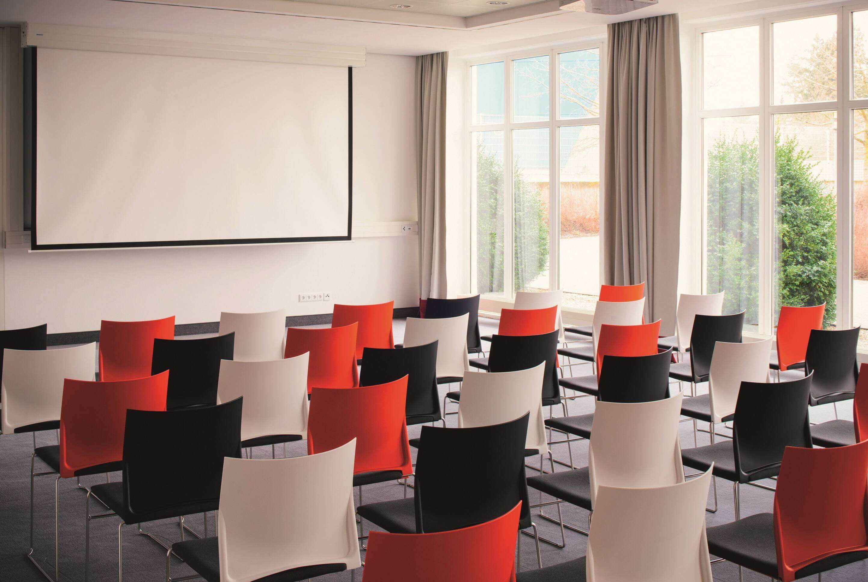 A conference room with rows of chairs, a projector screen, and large windows with curtains.