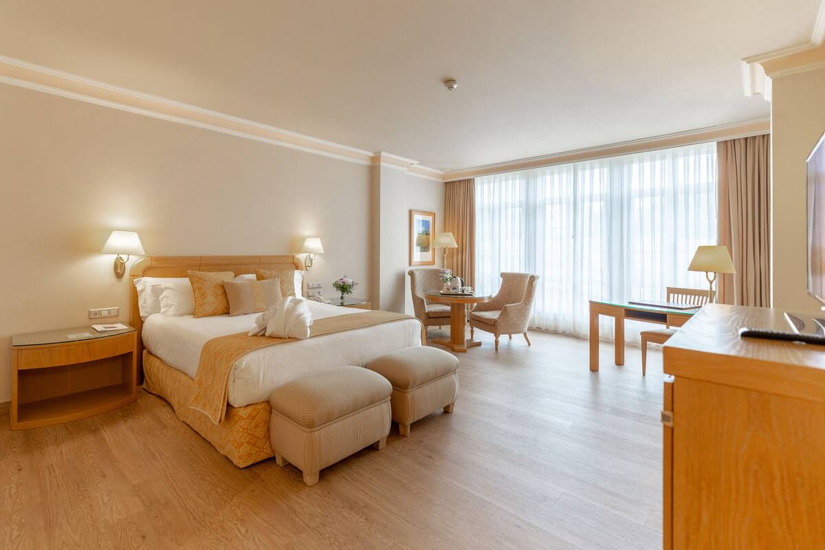 Een hotelkamer met een groot bed, twee nachtkastjes, een bureau met stoel, een televisie en een groot raam met gordijnen.
