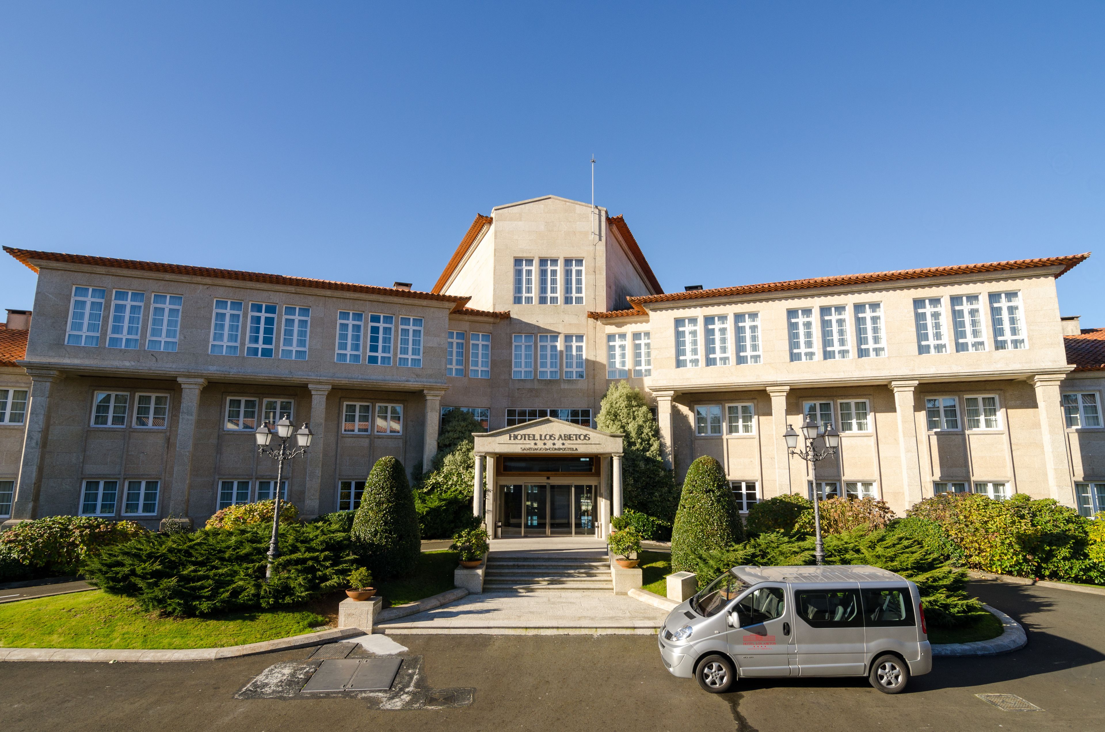 Un edificio grande de dos pisos con fachada simétrica, una entrada central con un letrero y un área de estacionamiento con una camioneta plateada.