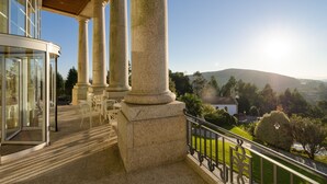 Un balcon orné de colonnes en pierre, une entrée avec une porte vitrée et une vue sur une vallée où l’on aperçoit une église et des arbres.