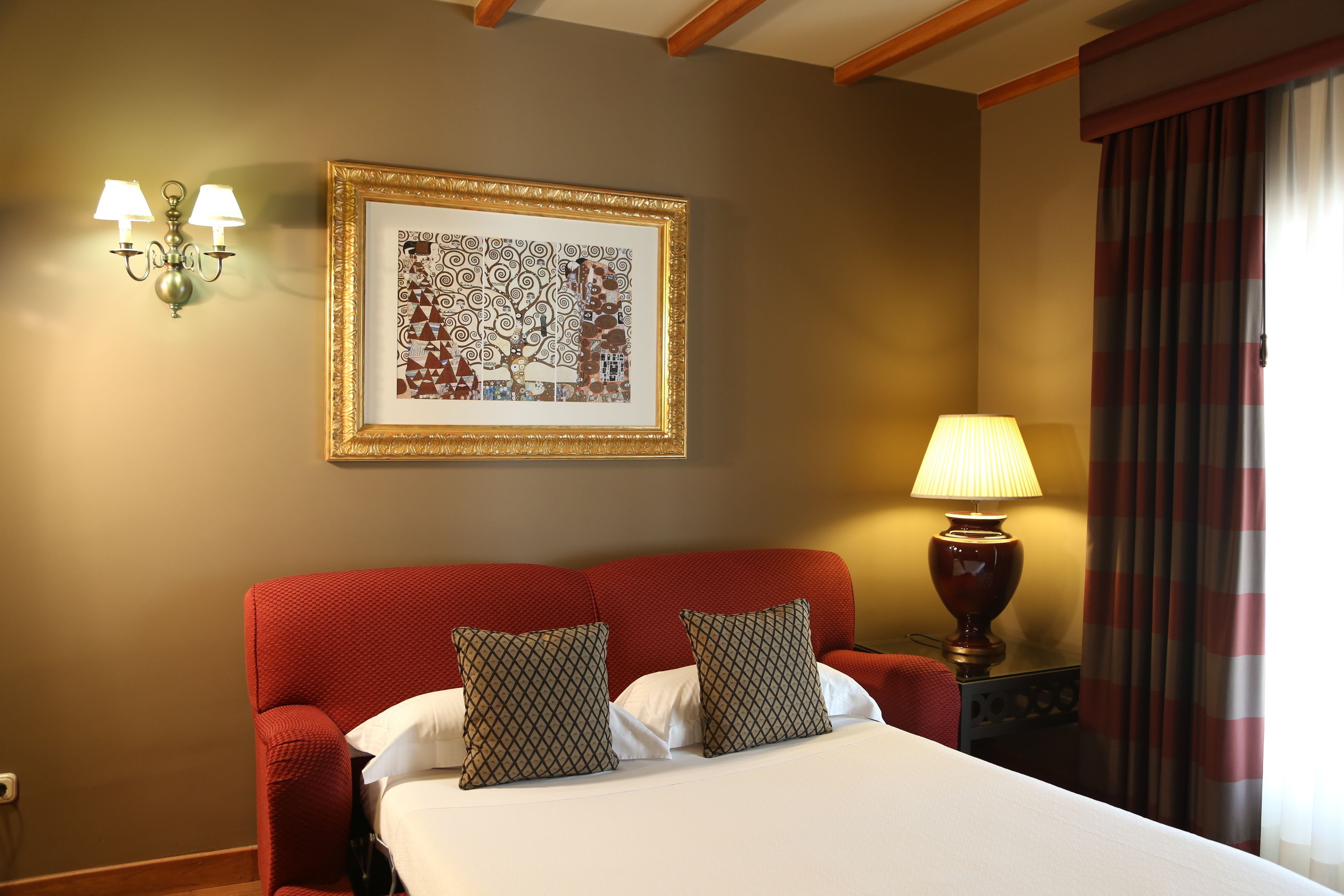 A hotel room with a large bed, two pillows, a framed artwork, a lamp, and a window with curtains.
