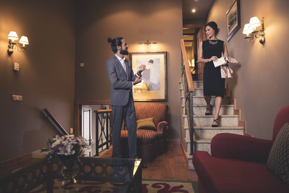 Een man en een vrouw staan in een hal met een trap, een ingelijst schilderij aan de muur en een rode bank.
