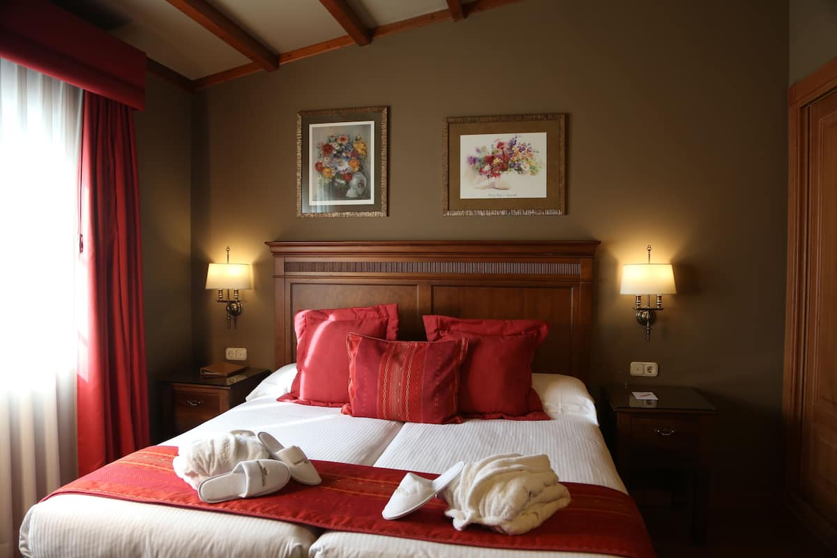 Een hotelkamer met een groot bed, rode kussens, twee wandlampen en twee florale foto's in lijsten.