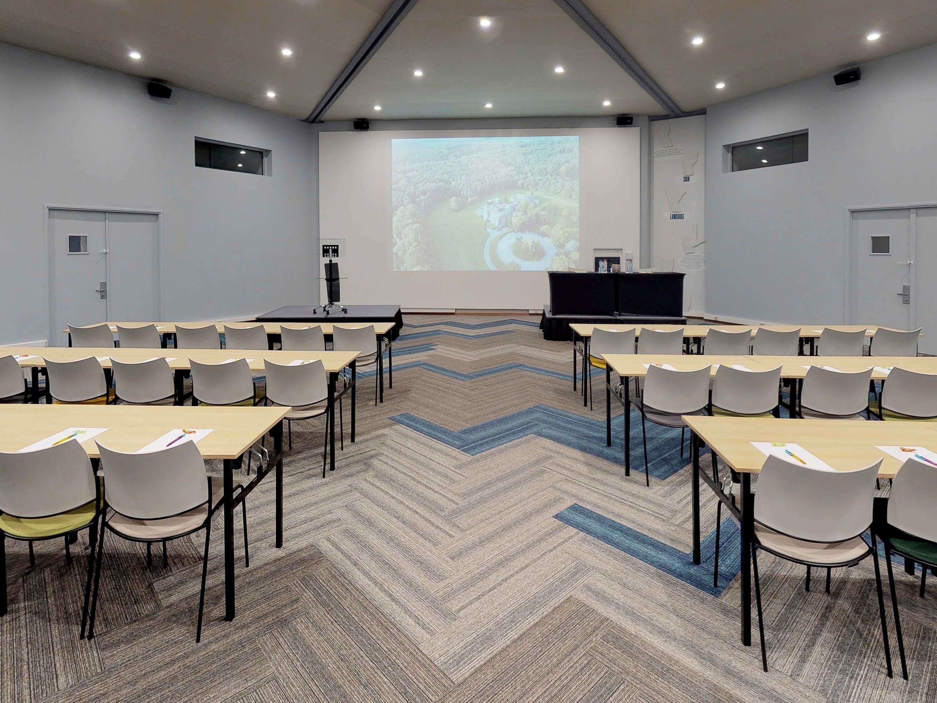 A conference room with a large screen, multiple tables, and chairs arranged for presentations.