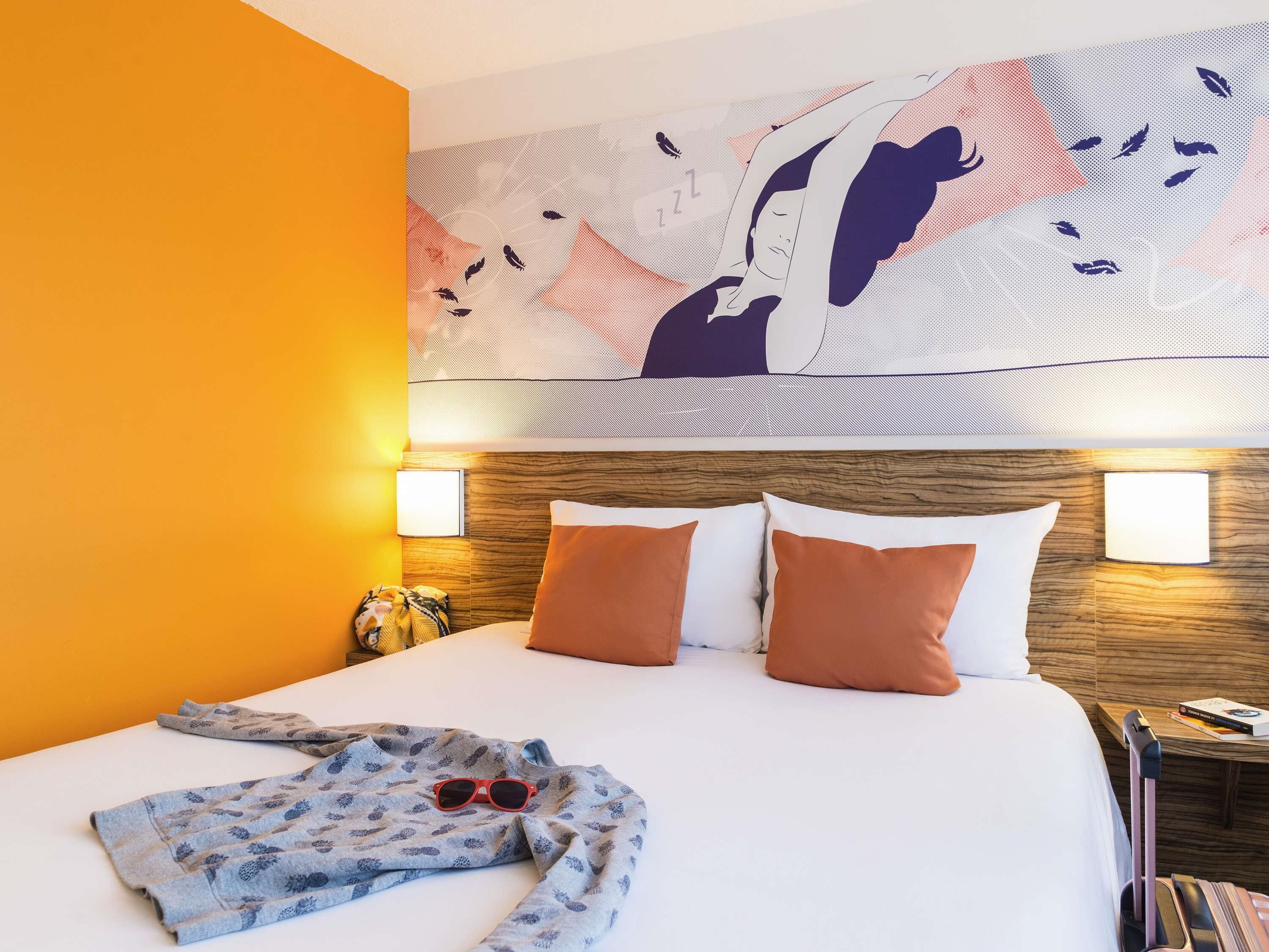 Una habitación de hotel con cama, almohadas, una colcha, unas gafas de sol, una mesita de noche y una obra de arte en la pared.