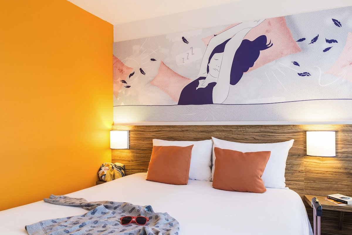 Ein Hotelzimmer mit Bett, Kissen, Decke, einer Sonnenbrille, einem Nachttisch und einem Wandbild.
