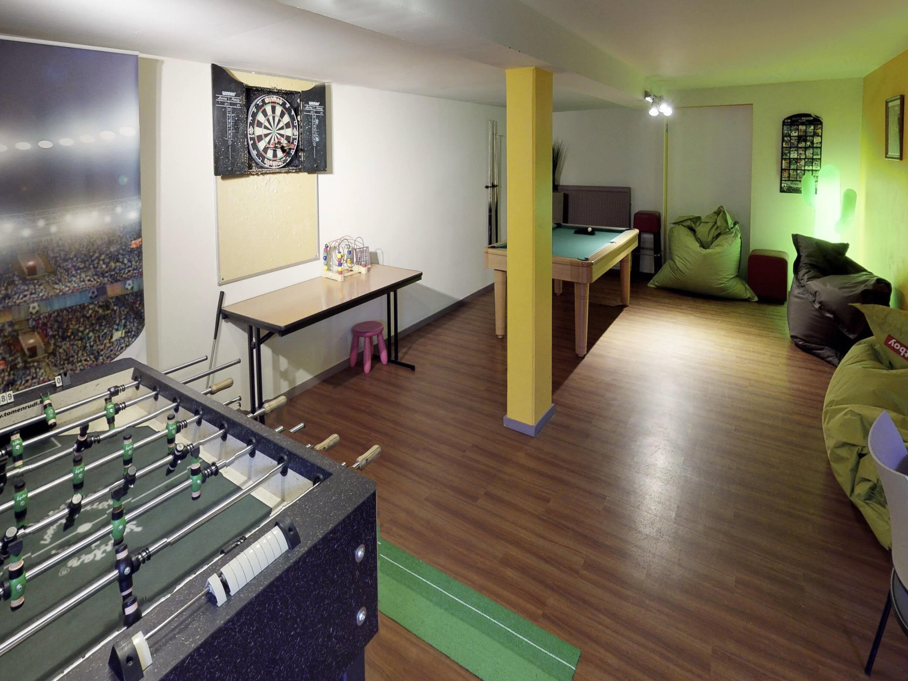 A room with a foosball table, a dartboard, a pool table, and a sofa.