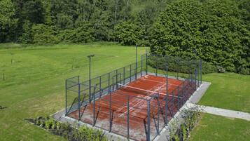 A tennis court surrounded by a metal fence, with a grassy field and trees in the background.