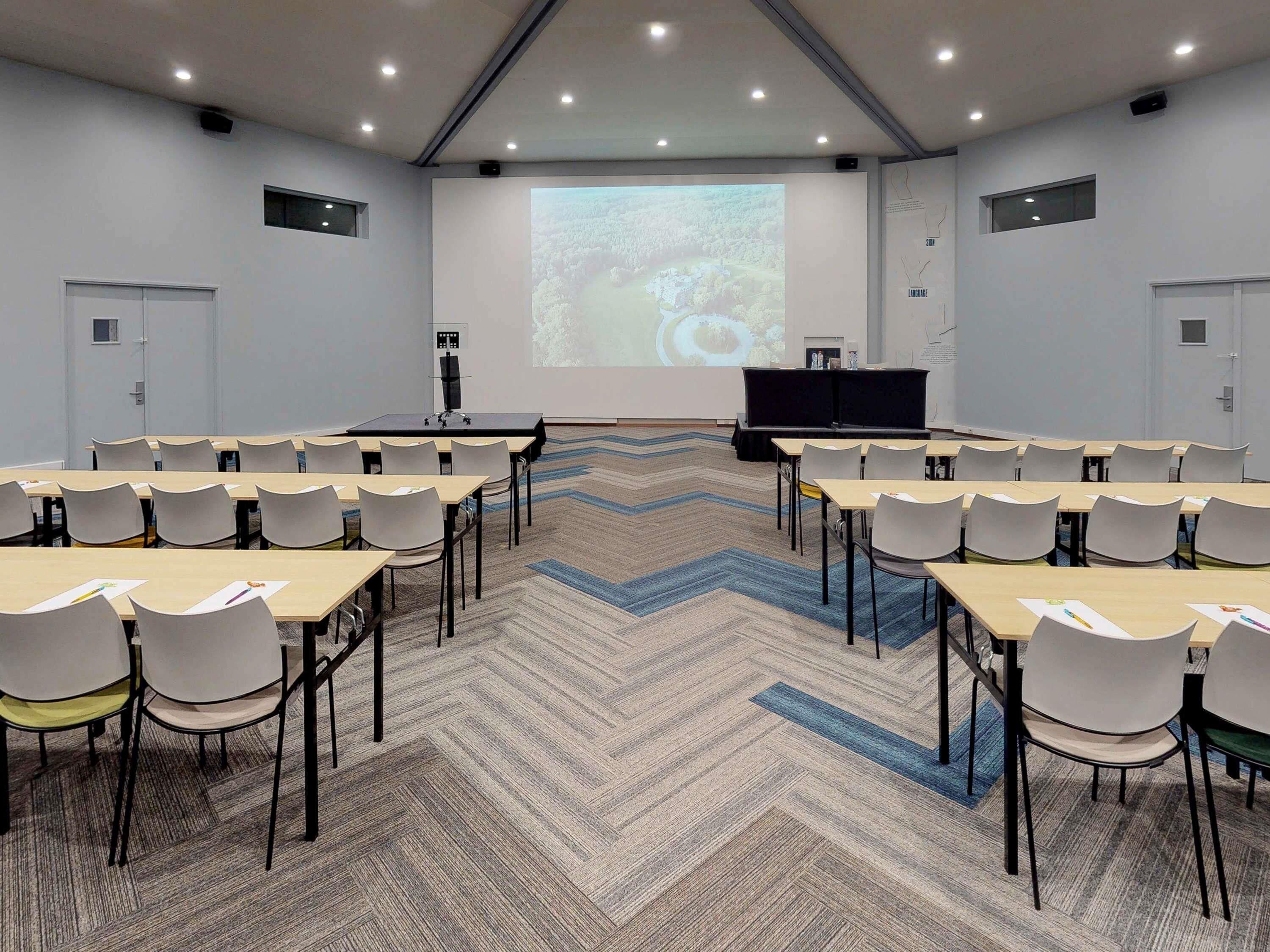 Un salón de conferencias con una pantalla grande, varias mesas y sillas dispuestas para presentaciones.