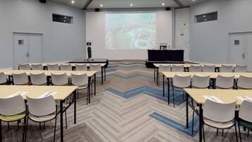 Un salón de conferencias con una pantalla grande, varias mesas y sillas dispuestas para presentaciones.