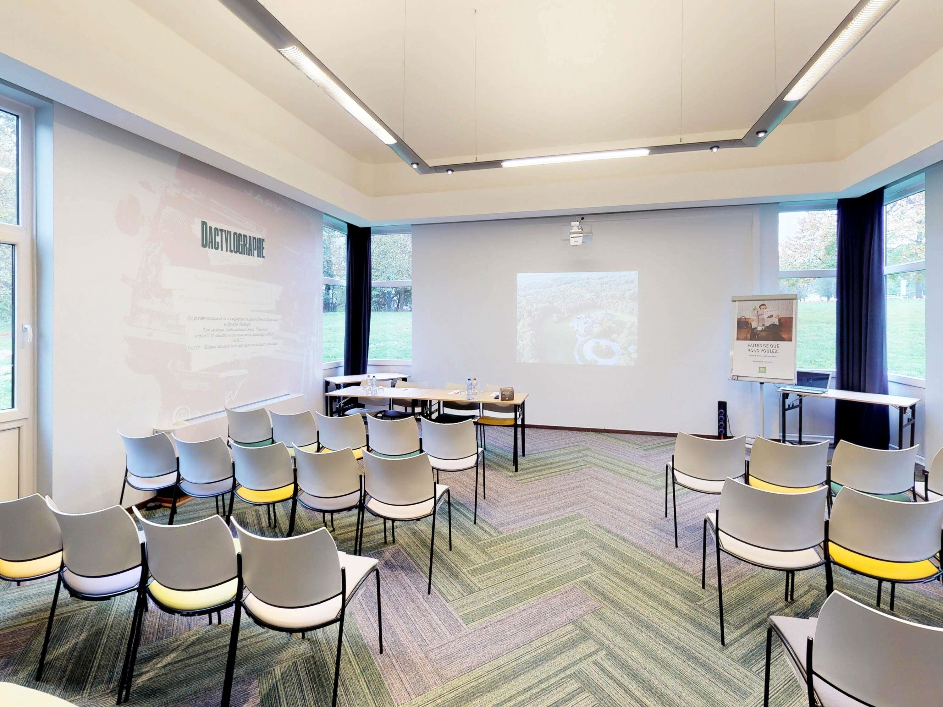 Sala de conferência com tela de projeção, cadeiras brancas, piso coberto por carpete e janelas amplas.