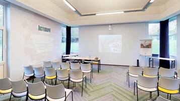 Sala de conferência com tela de projeção, cadeiras brancas, piso coberto por carpete e janelas amplas.