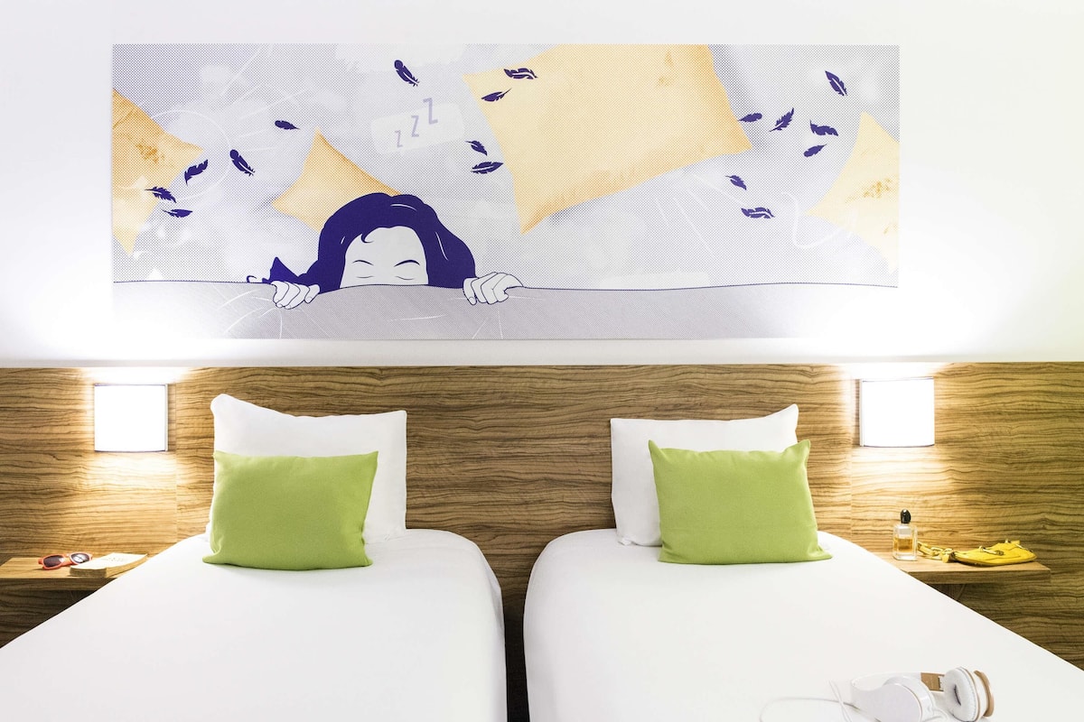 Ein Hotelzimmer mit zwei Betten, jedes mit einem grünen Kopfkissen, einem Holzkopfbrett und einem Wandbild darüber.