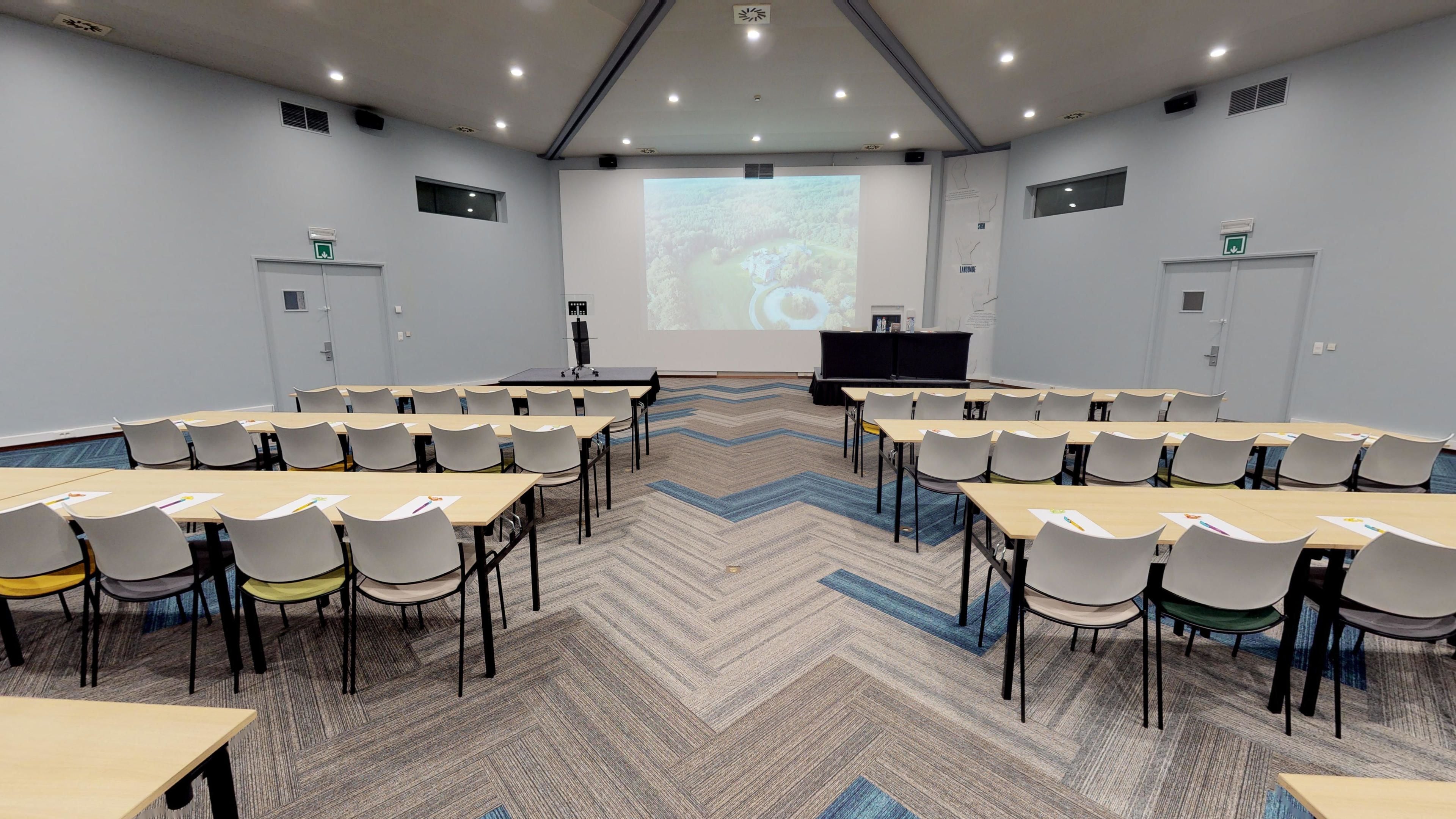 A conference room with a large screen at the front, multiple tables and chairs arranged in rows, and a carpeted floor.