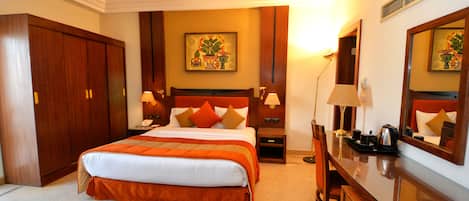 A hotel room with a large bed, a desk, and a chair. There is a painting on the wall and a lamp on the desk.