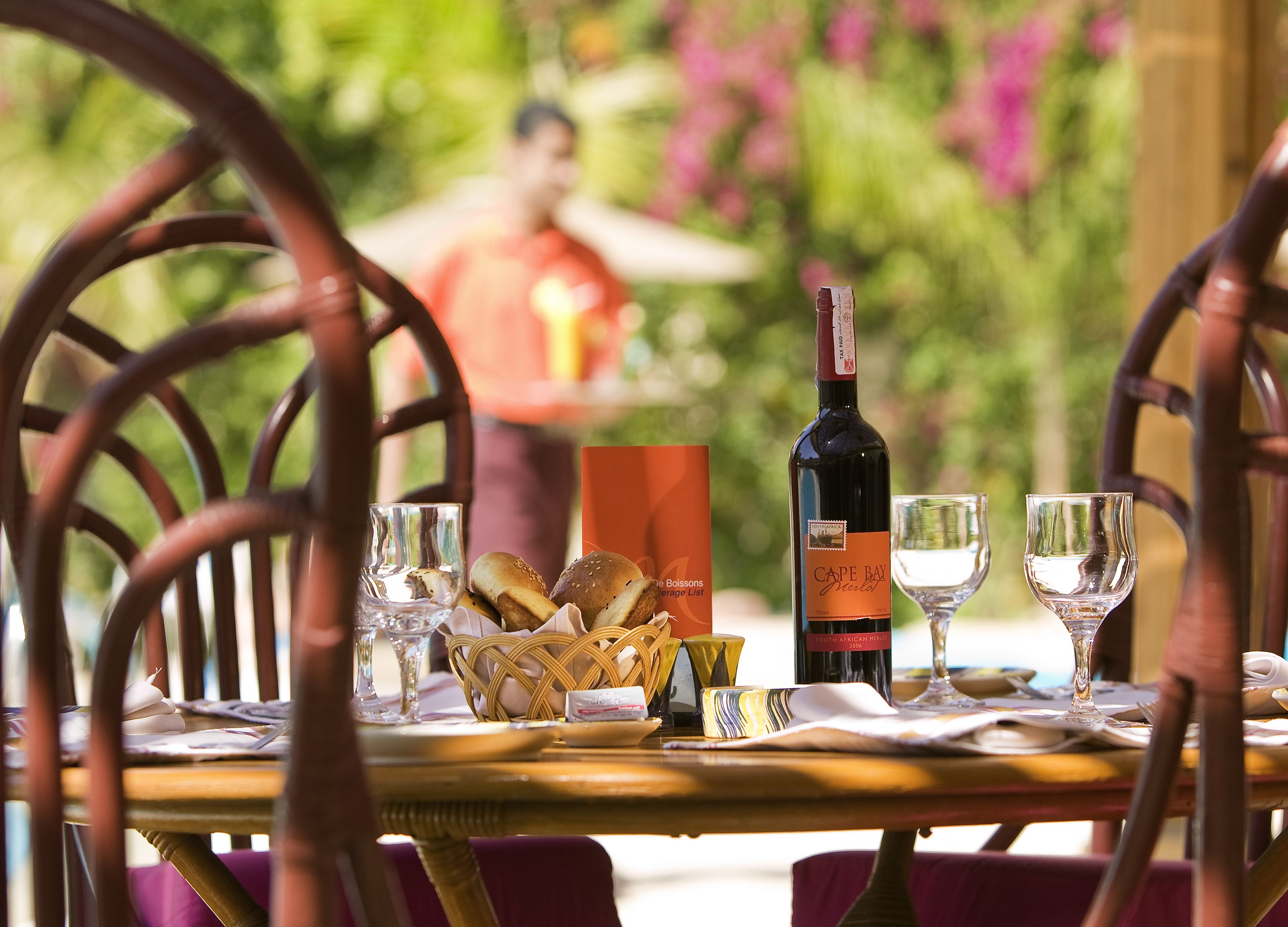 A dining table set with a bottle of Cape Bay Merlot, two glasses, a basket of bread, and a menu.