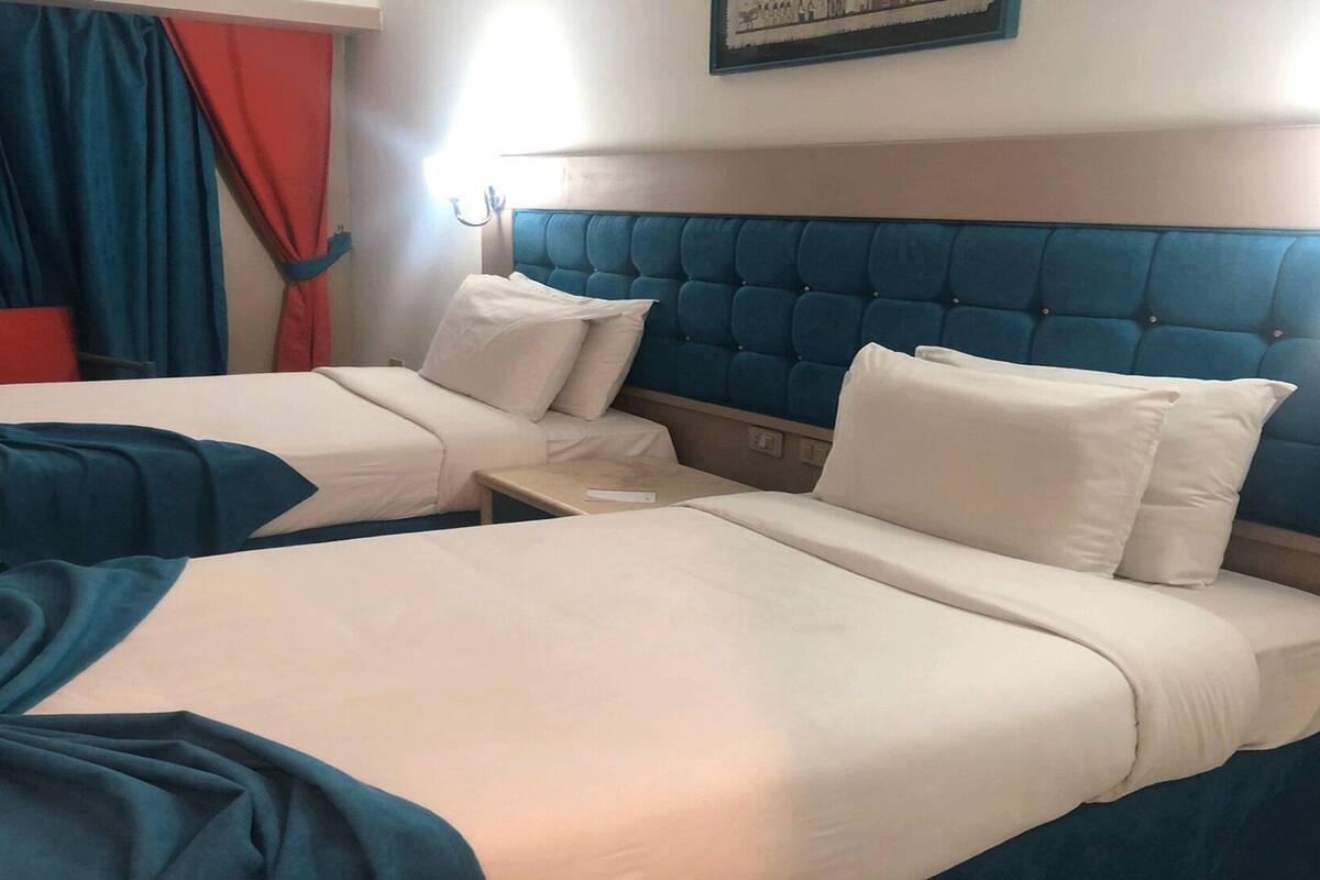 A hotel room with two beds, each with white bedding and blue headboards. There is a nightstand between the beds, and a red curtain is visible on the left side.