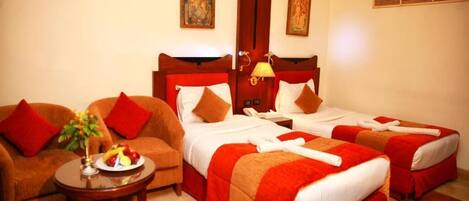 A hotel room with two beds, a sofa, a small table with a fruit plate, and artwork on the walls.