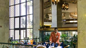 Une salle à manger avec une table mise pour le repas, un homme en costume qui prépare de la nourriture, et une vue sur l’extérieur à travers de grandes fenêtres.