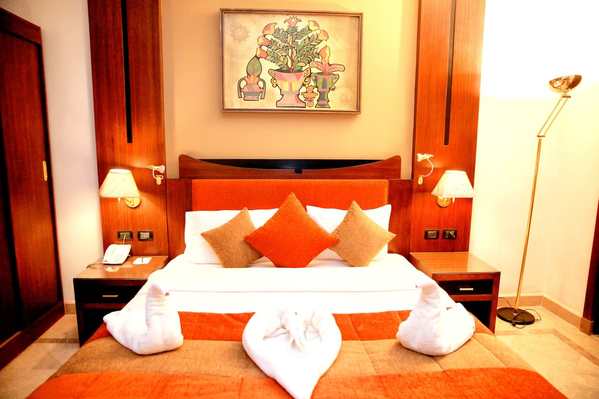 A hotel room with a bed, two bedside tables, a nightstand, a lamp, a painting on the wall, and a floor lamp.