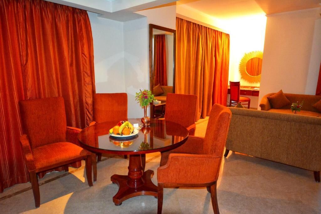 Een kamer met een ronde glazen tafel omringd door vier stoelen, een bank en een eettafel met een fruitschaal.
