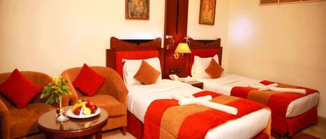 A hotel room with two beds, a sofa, a small table with a fruit plate, and two paintings on the wall.